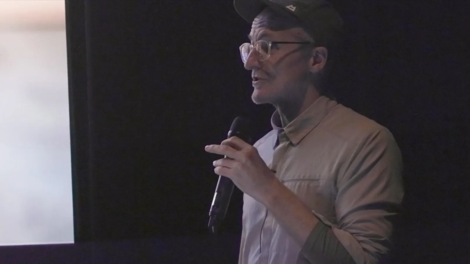 Jhave speaking into a hand-held microphone in the dark cinema, beside the edge of the lit screen.